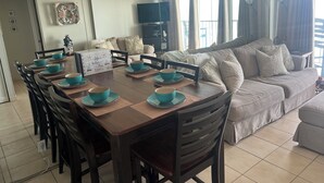 A dining area with a wooden table set for four, surrounded by chairs. A sofa and a television are visible in the background. - Relax By The Sea With Infinity Ocean Views (Galveston)