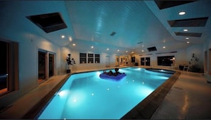 A large indoor swimming pool with a blue water feature, surrounded by walls with windows and doors. - Heated Indoor Pool and Spa - Golf Course View (Mashpee)