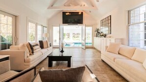 A spacious living room with a large window, a sofa, a coffee table, and a television. - Heated Indoor Pool and Spa - Golf Course View (Mashpee)