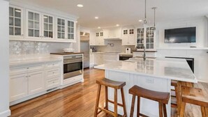 A modern kitchen with white cabinetry, a central island, wooden stools, and a large TV mounted on the wall. - Heated Indoor Pool and Spa - Golf Course View (Mashpee)