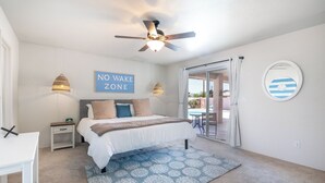 A bedroom with a bed, a nightstand, a desk, a ceiling fan, a 'NO WAKE ZONE' sign, and a view of the outside. - STOCKED Pool Home, RV & Boat parking, Game room. Sleeps 13 (Lake Havasu City)