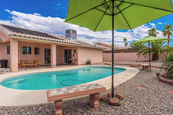 A swimming pool with a green umbrella, a patio with a table and chairs, and a building with a tiled roof. - STOCKED Pool Home, RV & Boat parking, Game room. Sleeps 13 (Lake Havasu City)