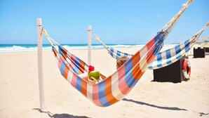 Escena de playa con una hamaca atada entre dos postes, un flotador salvavidas y un cielo azul despejado.