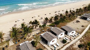 Un complejo turístico en la playa con cabañas de techo de paja, sillones de sol y palmeras.