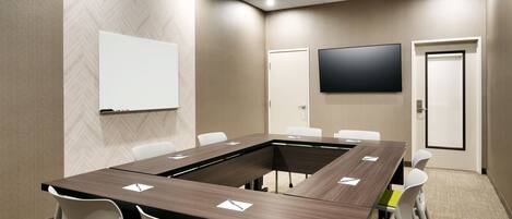 A conference room with a long table, white chairs, a whiteboard, and a flat-screen TV on the wall.