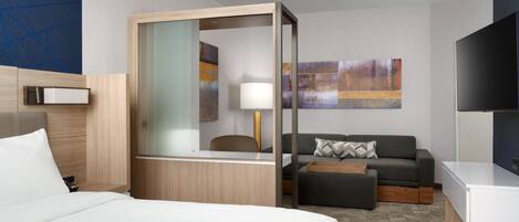 A modern hotel room with a bed, a desk, a sofa, a TV, and abstract art on the wall.
