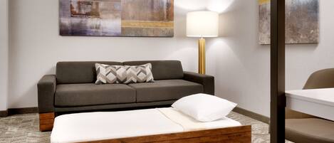 A hotel room with a sofa bed, a bedside table, a lamp, and abstract wall art.