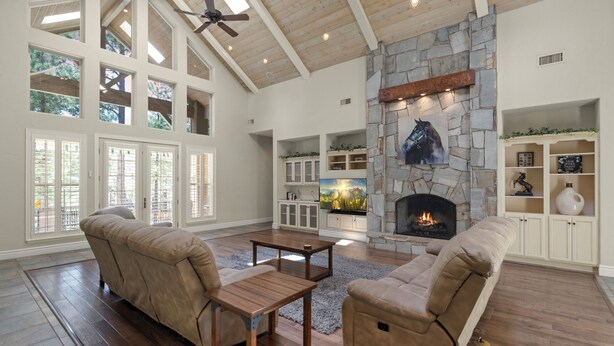 A spacious living room with a stone fireplace, wooden flooring, and a large window with multiple panes. - Lone Oak (Flagstaff)