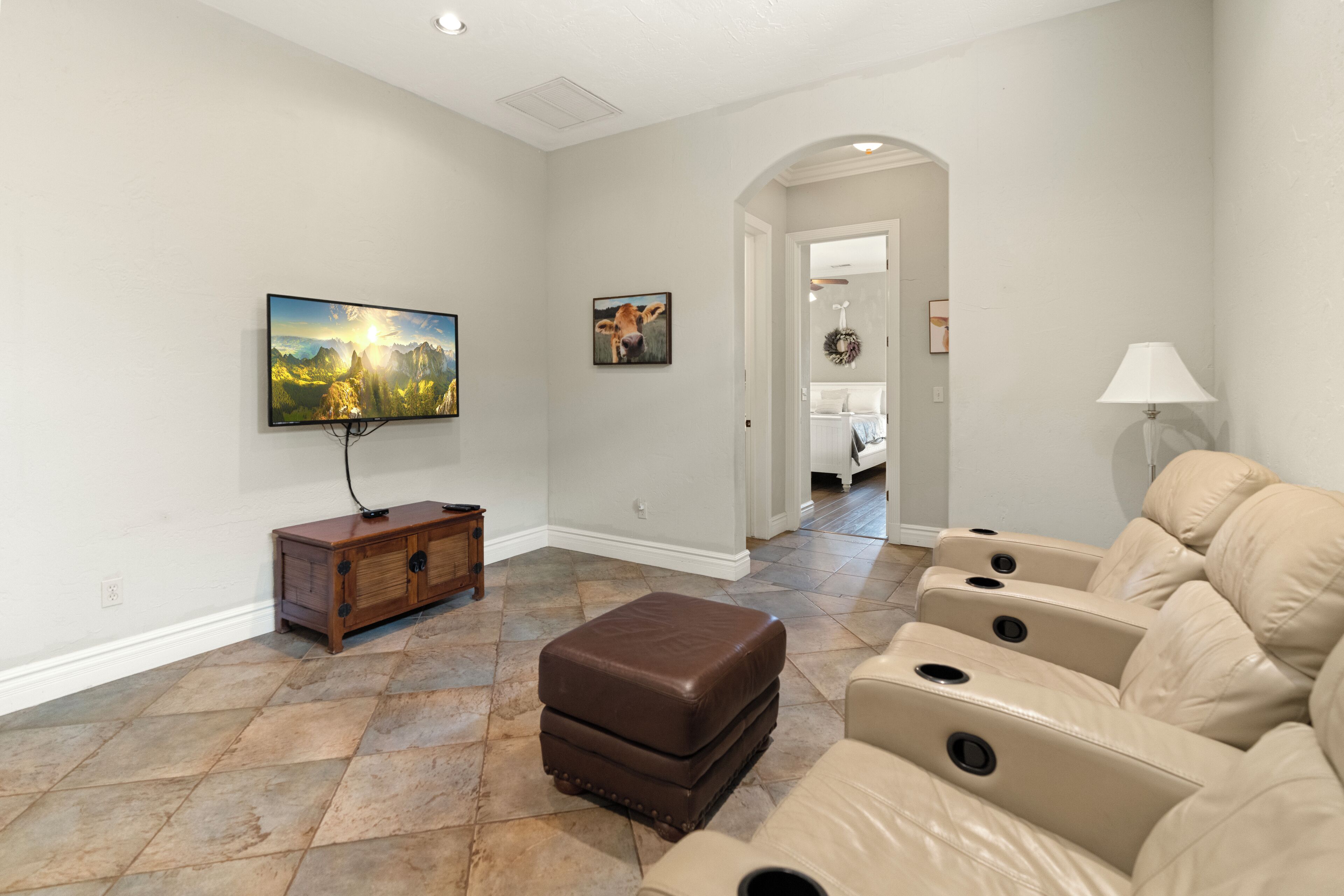 A living room with a flat-screen TV, a brown ottoman, beige leather sofas, and a stone tile floor.