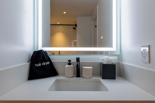 Un baño con un lavabo blanco, un espejo, una bolsa para secador de pelo, un dispensador de jabón y una caja de pañuelos.