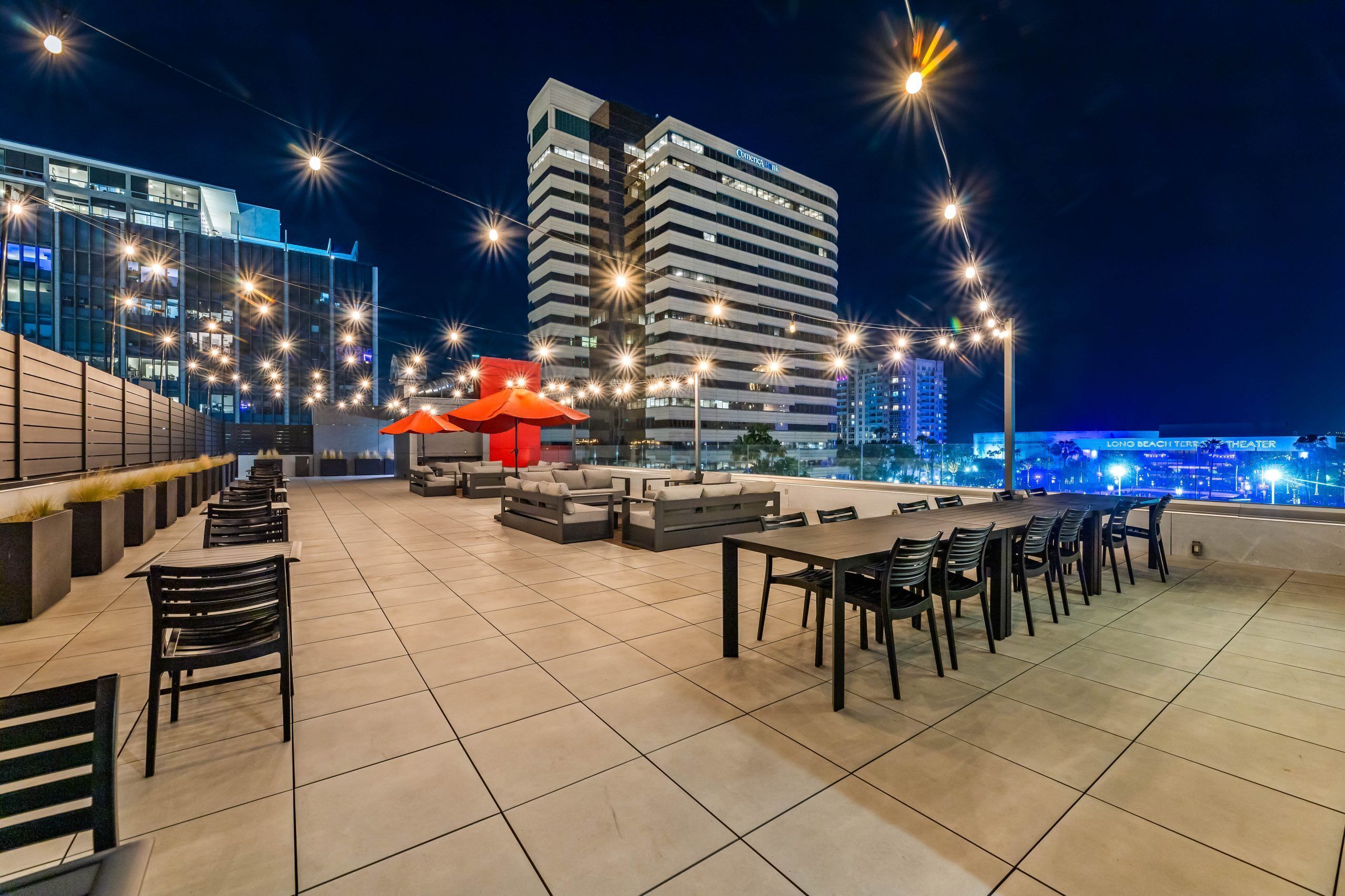 A rooftop terrace with outdoor seating, a red umbrella, and a view of a city skyline at night.
