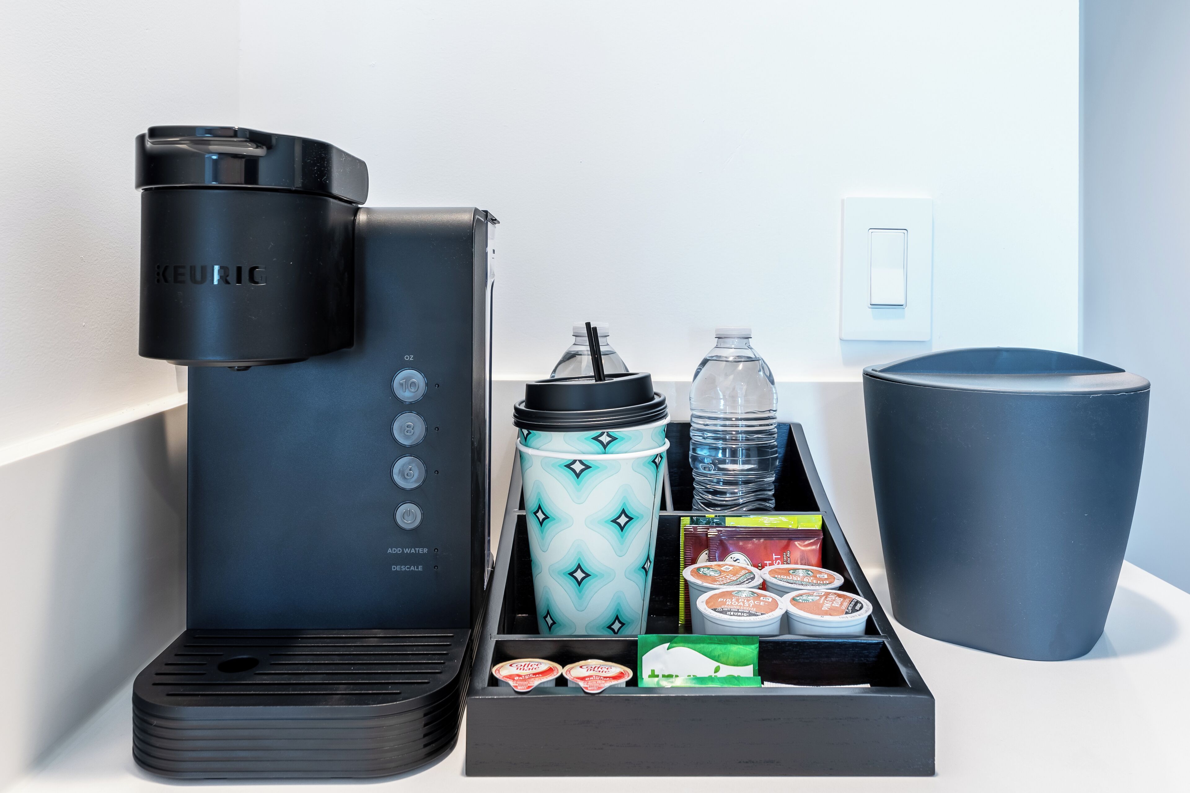 Una cafetera, una botella de agua, un vaso tipo tumbler y un recipiente con vasos de yogur sobre una superficie blanca.