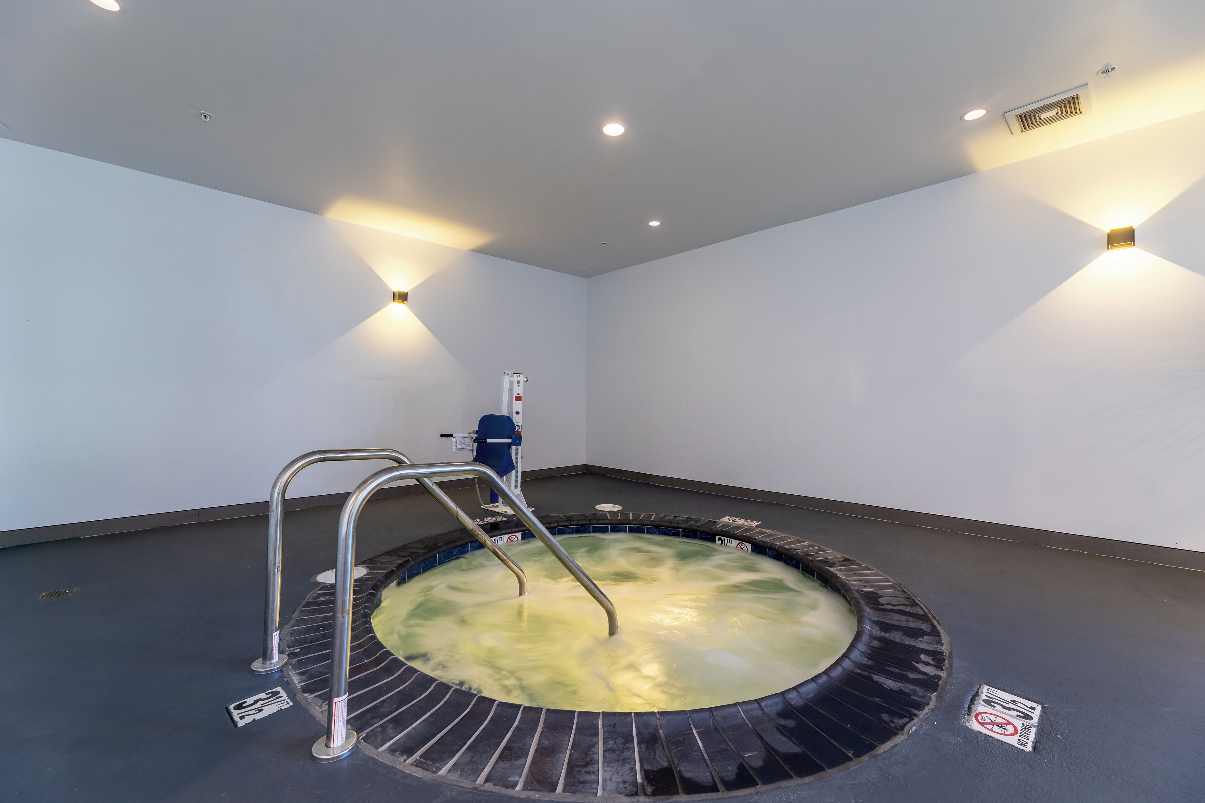 Un jacuzzi con barandas de metal, rodeado de un piso de baldosas, con un panel de control y una lámpara fijada a la pared.