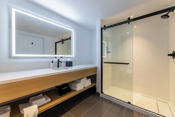 Un baño moderno con cabina de ducha de vidrio, un espejo grande con iluminación integrada y toallas ordenadas en un estante de madera.
