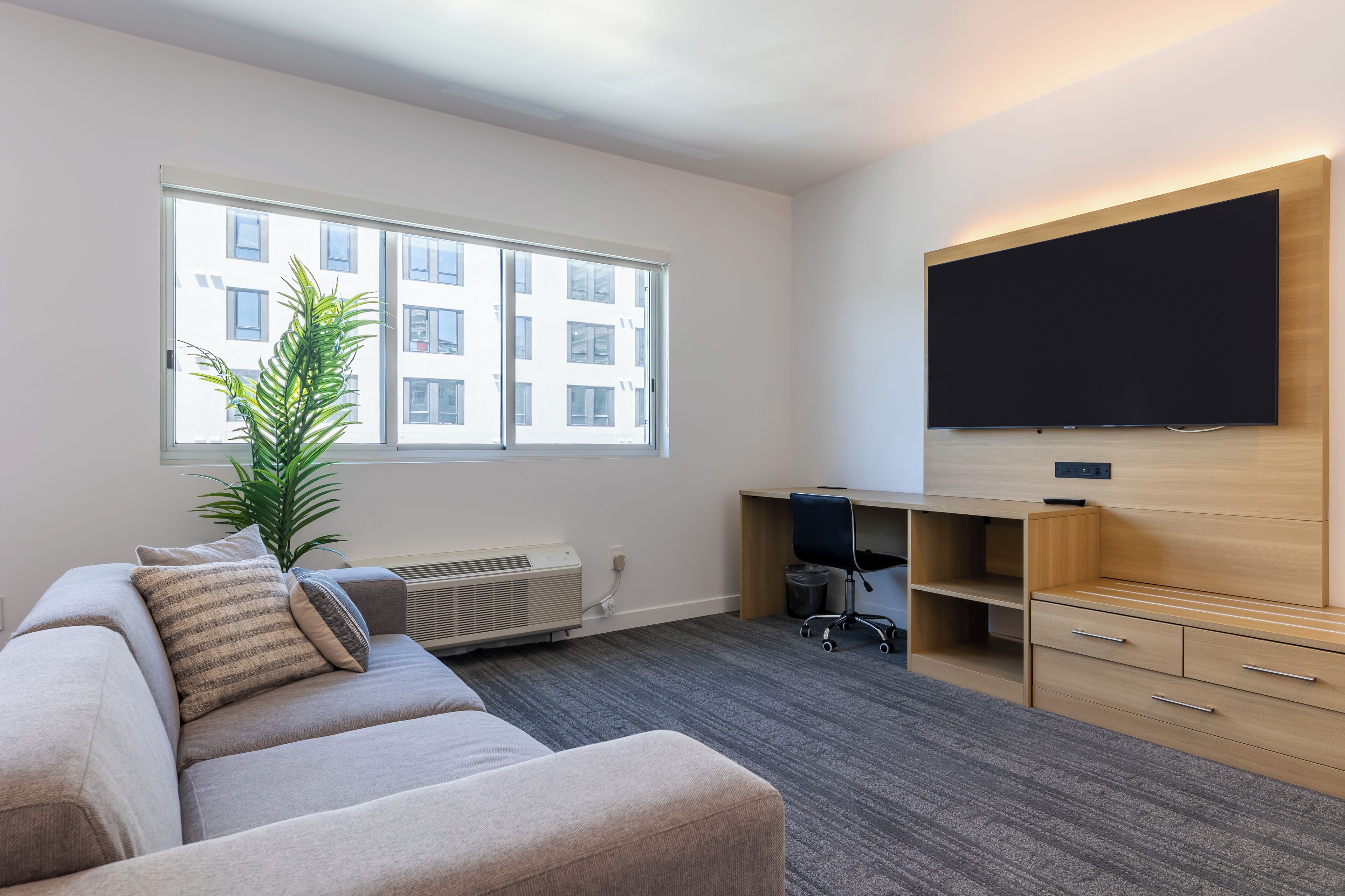 A modern living room with a sofa, a flat-screen TV mounted on the wall, a desk with a chair, and a large window with a view of buildings.