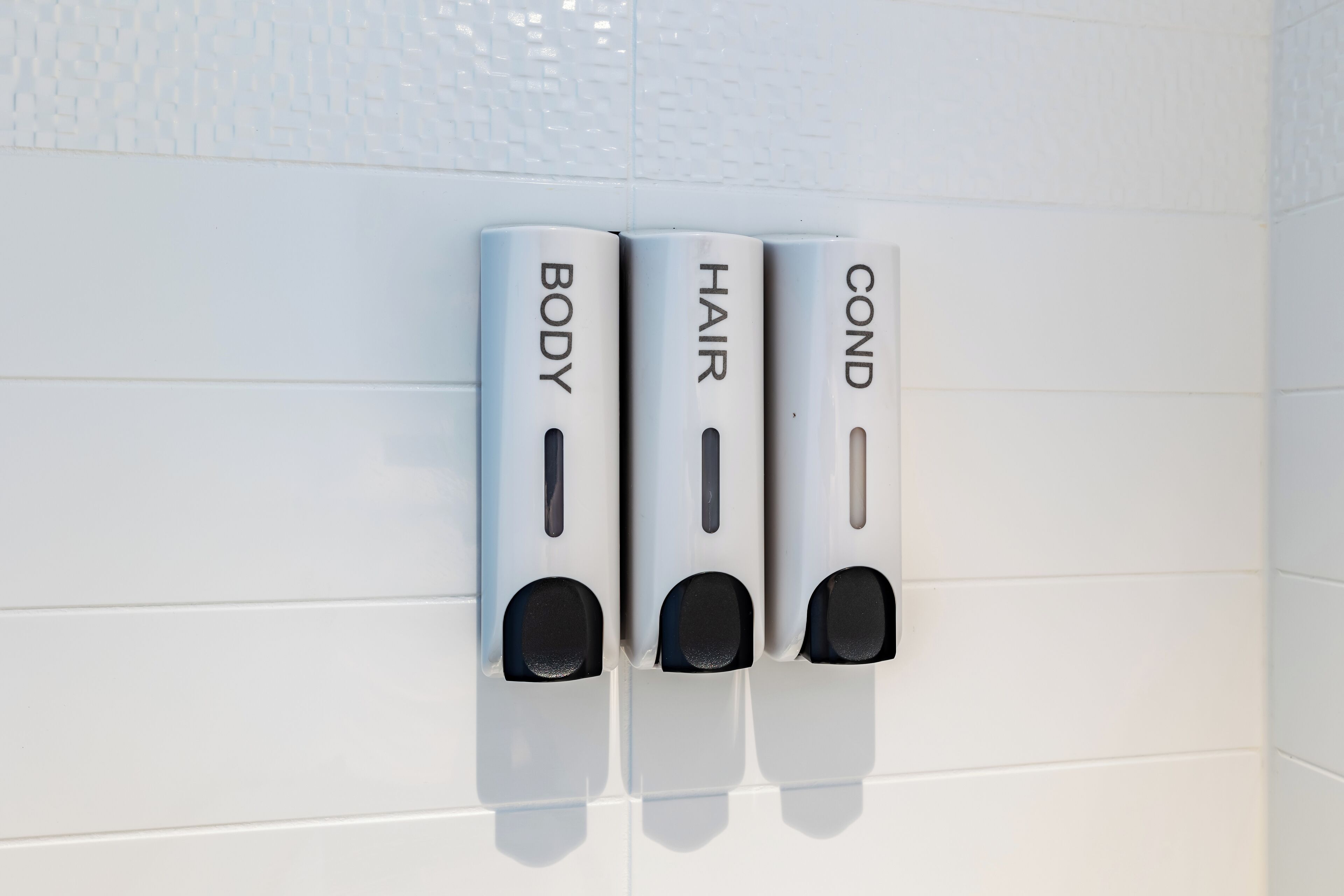 Three hand soap dispensers labeled BODY, HAIR, and COND on a white tiled wall.