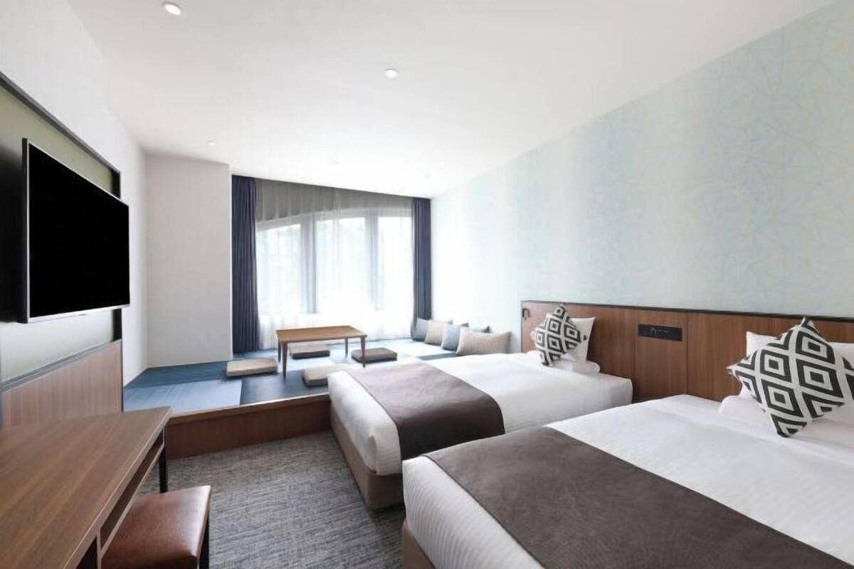 A hotel room with two beds, a flat-screen TV, a desk, and a window with curtains.