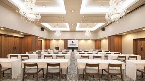 Un salón de conferencias con filas de mesas y sillas, candelabros y una pantalla de presentación.