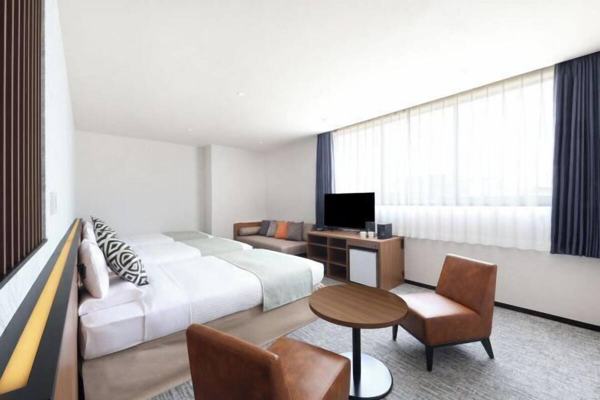 A modern hotel room with a bed, sofa, armchair, TV, and a small desk.