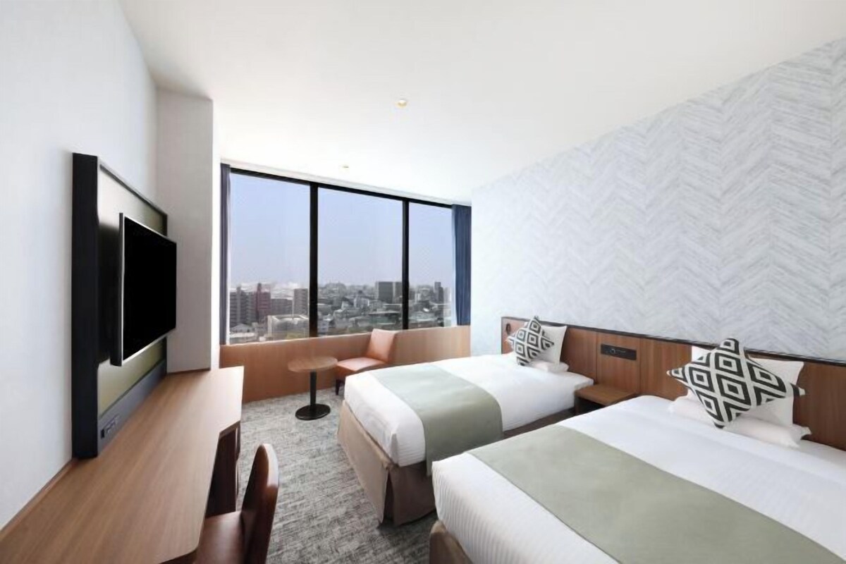 A modern hotel room with two beds, a flat-screen TV, a city view, and a herringbone-patterned wall.