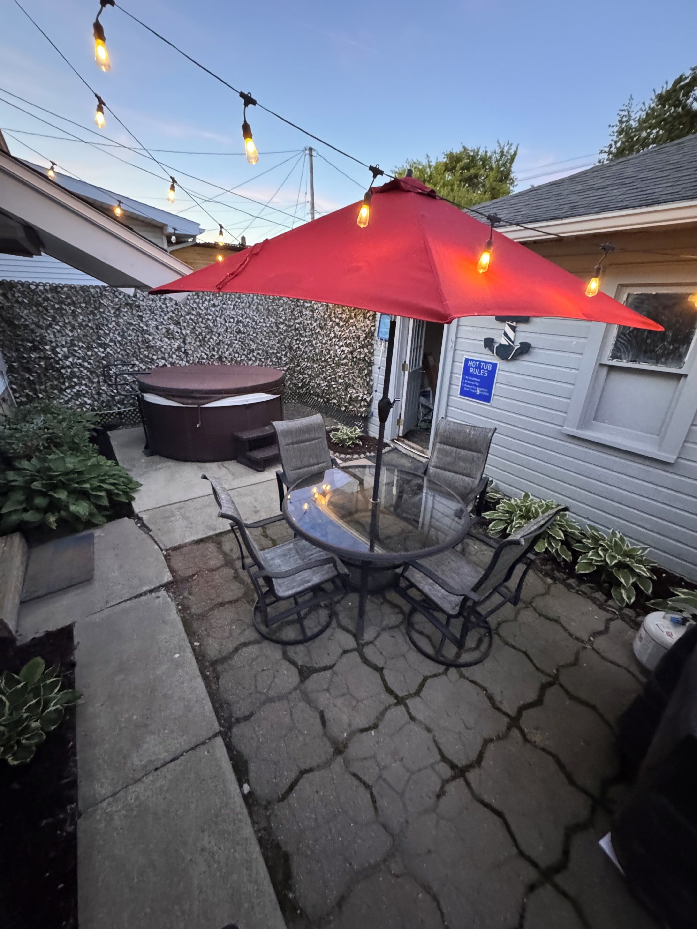 Un patio al aire libre con una mesa y sillas de vidrio, un paraguas rojo, luces colgantes y una zona de hidromasaje.