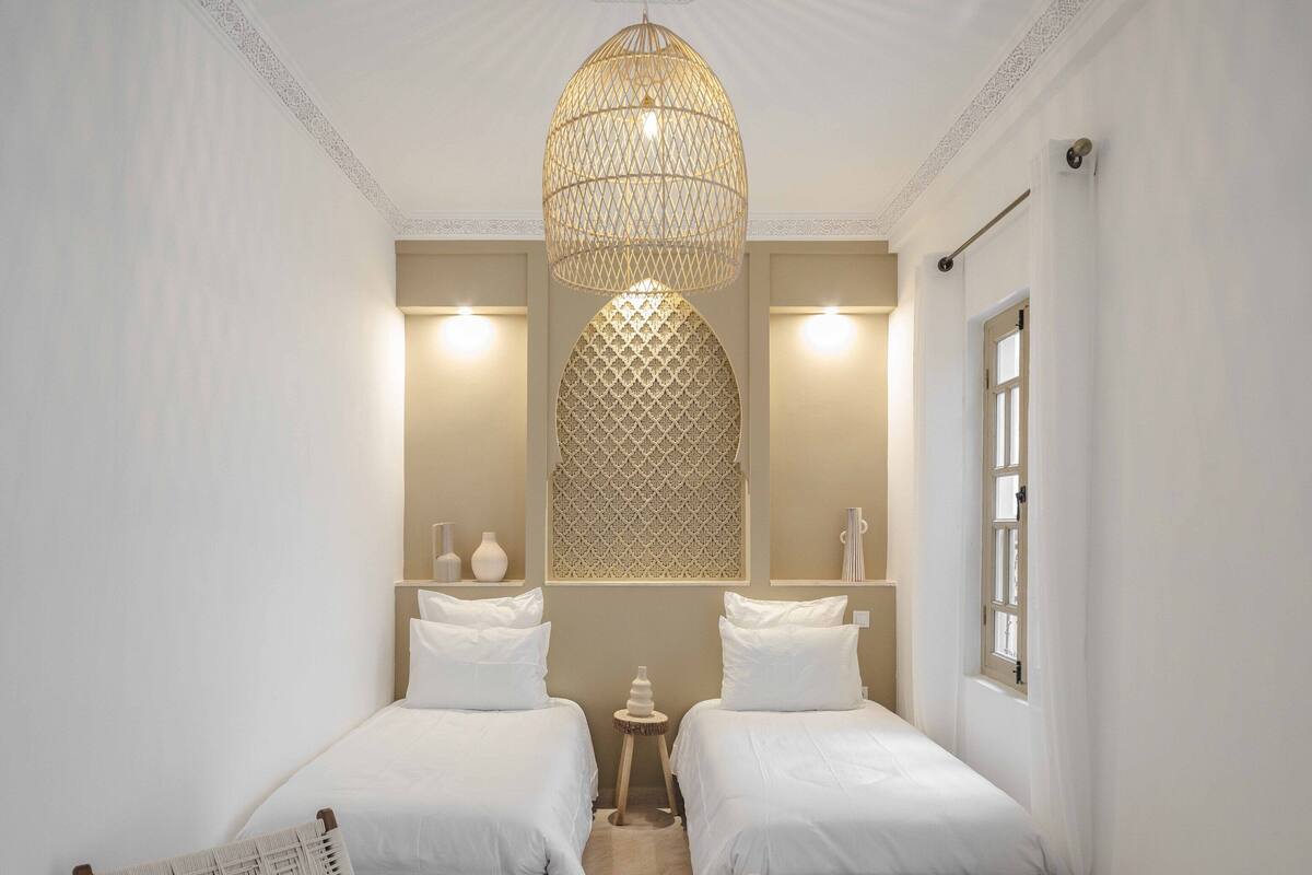 A hotel room with two beds, a woven chair, a small table with a candle, and a decorative ceiling light.
