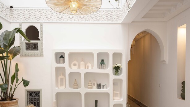 Un interior moderno con una mesa redonda de mármol, sillas blancas, una estantería decorativa con diversos objetos y una lámpara colgante.