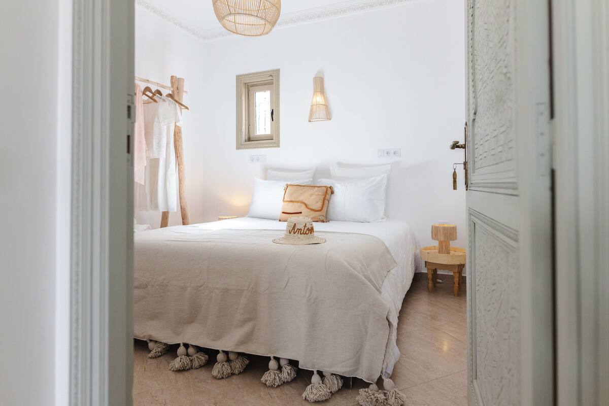 A bedroom with a bed, a bedside table, a hanging light, and a window.