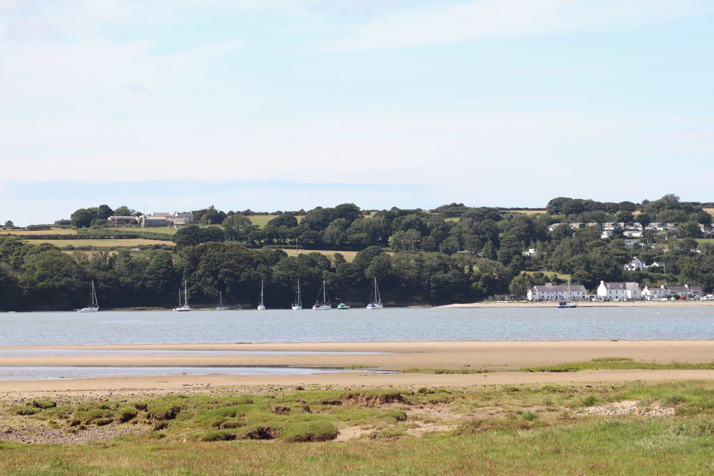 A coastal landscape with a sandy beach, sailboats, and a cluster of houses on a hill.