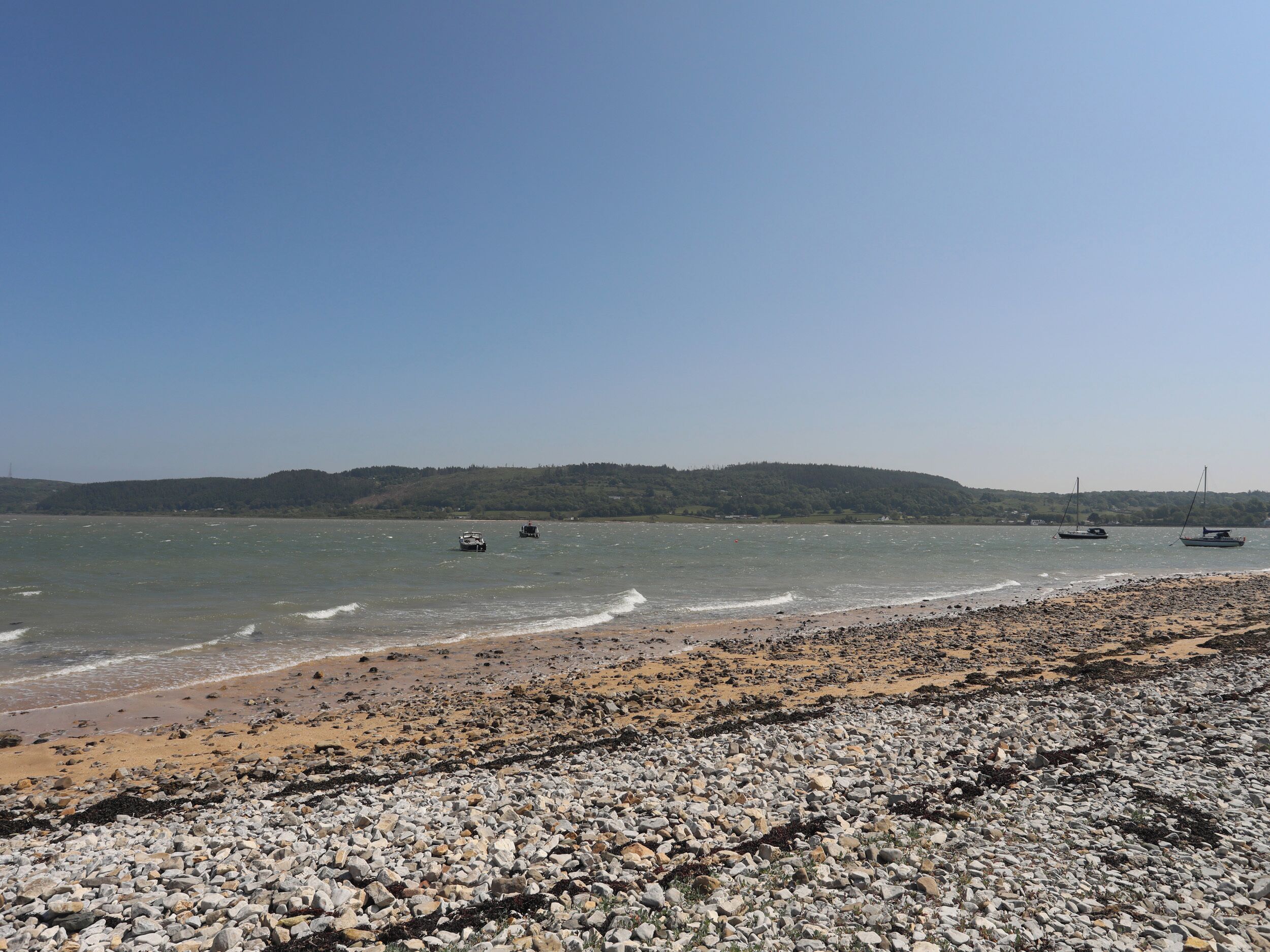 A pebbly beach with boats on the water and a forested hill in the background.