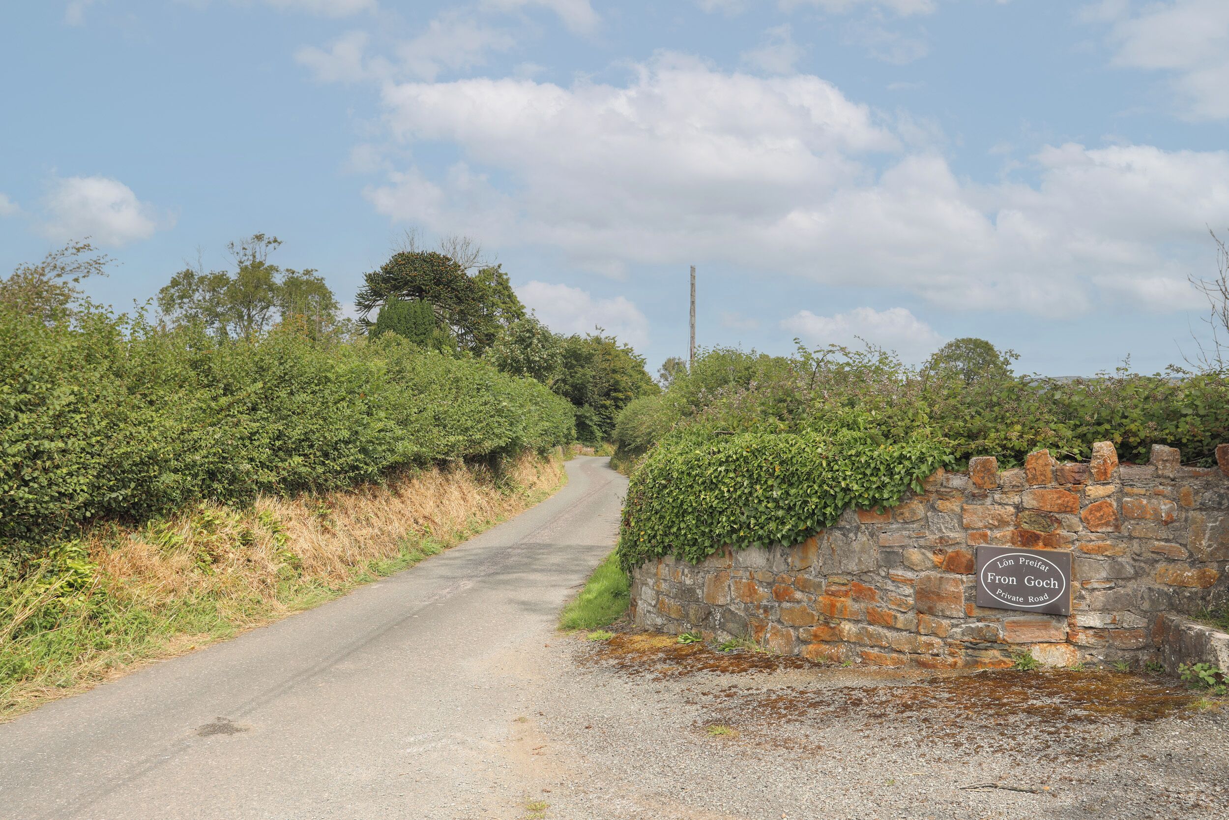 A rural road with a stone wall and a sign indicating 'Lon Profat Fran Goch Private Road'.