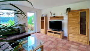 Un salon spacieux avec une table basse en verre, un canapé confortable, un téléviseur, des meubles en bois et une grande fenêtre donnant sur un paysage verdoyant.