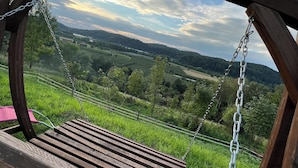 Une balançoire en bois avec des chaînes métalliques, d’où l’on peut admirer un paysage verdoyant et luxuriant, avec des champs et des arbres sous un ciel partiellement nuageux.