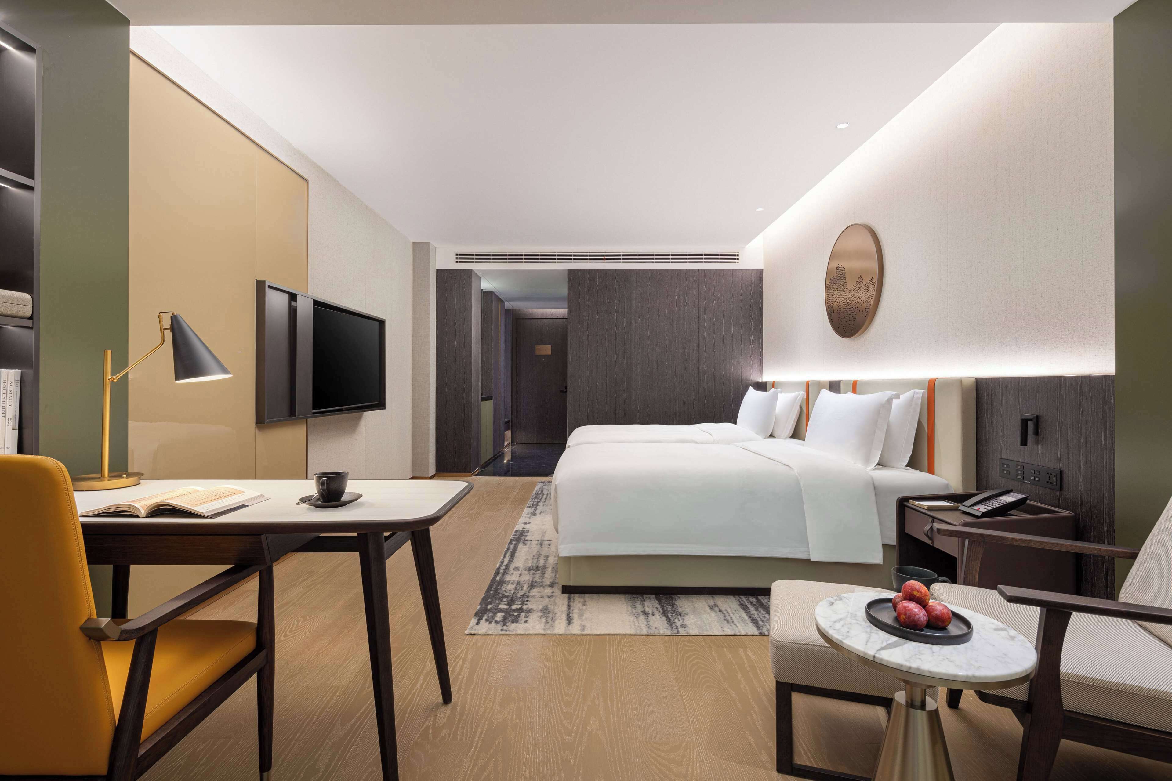 Une chambre d’hôtel moderne équipée d’un lit, d’un bureau avec une lampe, d’une chaise, d’une petite table avec des fruits et d’un téléviseur fixé au mur.