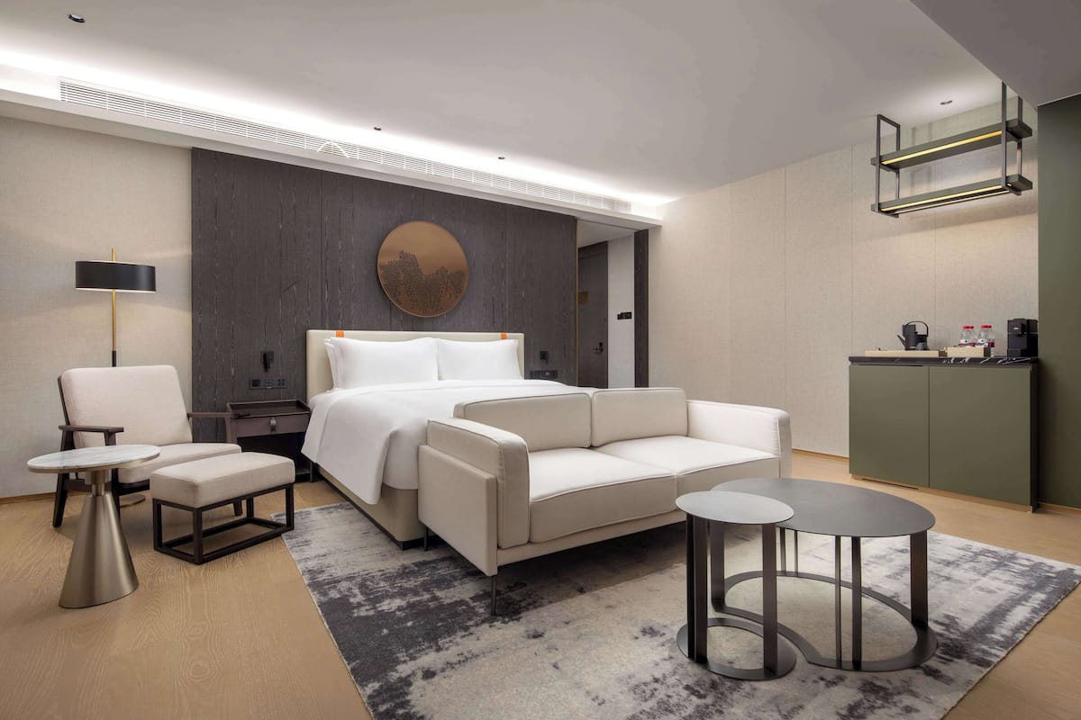Ein modernes Hotelzimmer mit Bett, zwei Sesseln, einem kleinen Tisch, einem runden Couchtisch, einem Nachttisch, einer Lampe, einem wandmontierten Spiegel und einer Küchenzeile.