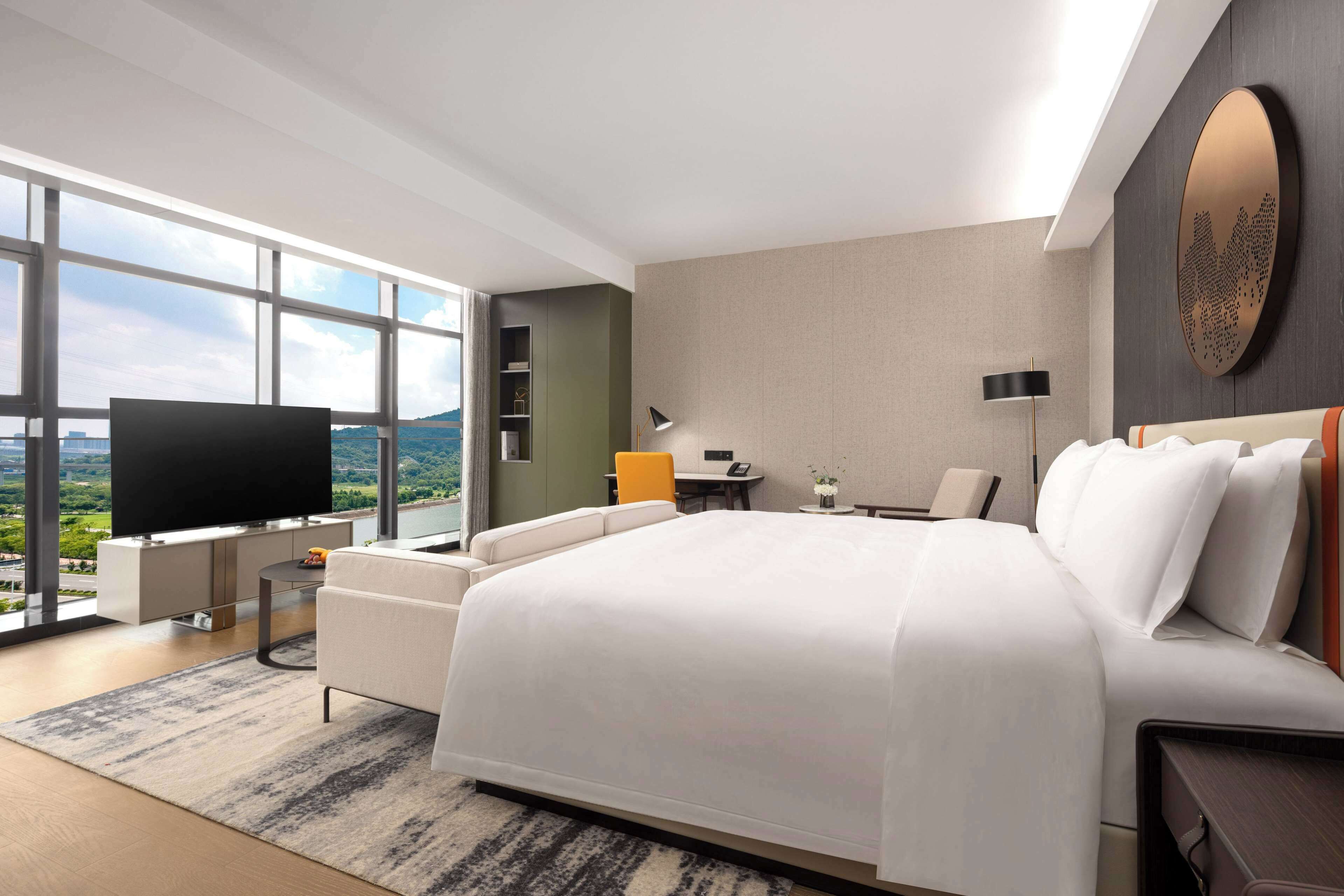 A modern hotel room with a large bed, a flat-screen TV, a desk with a chair, and a view of the outdoors through large windows.