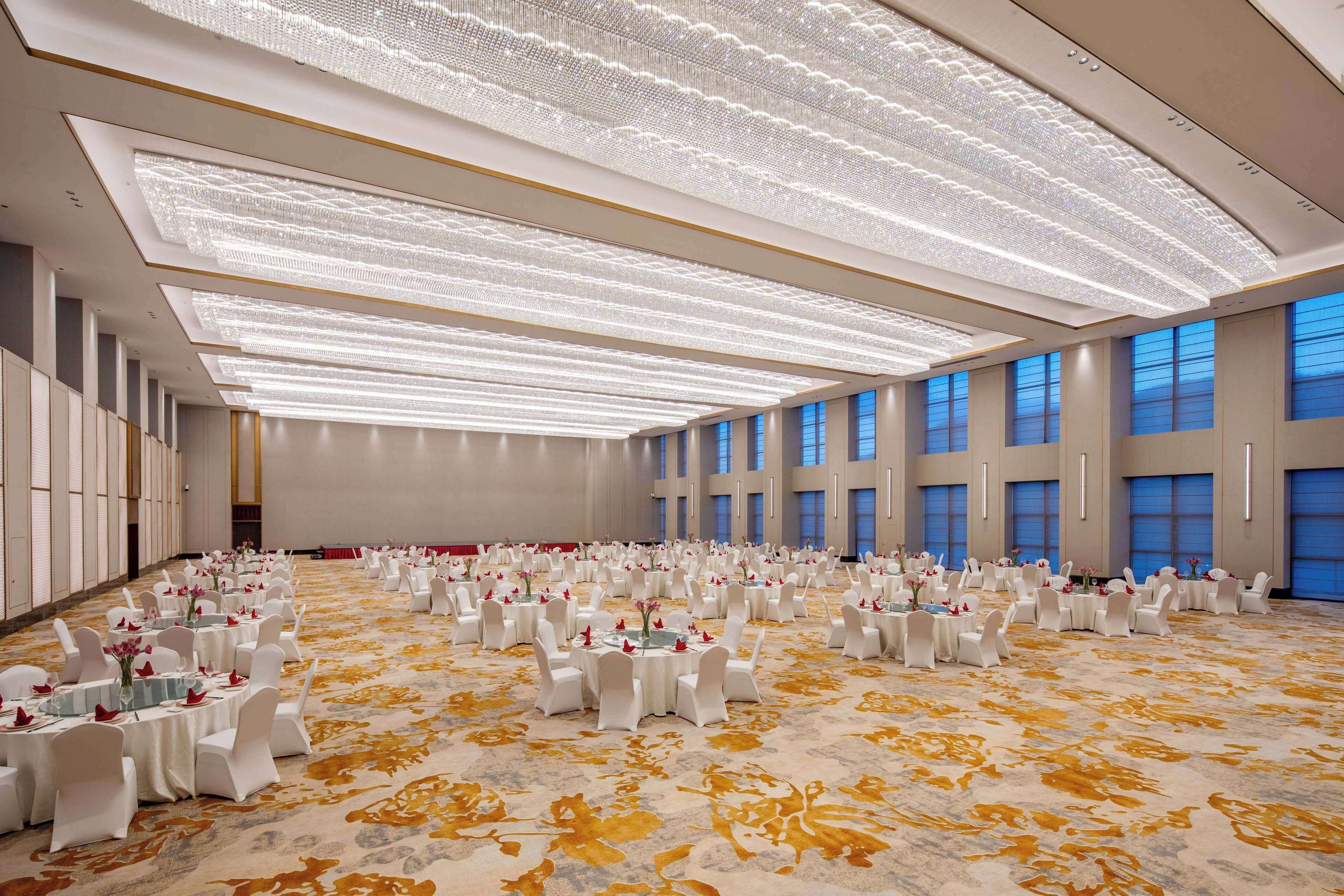A large, well-lit banquet hall with rows of round tables, each set with white chairs and floral centerpieces.