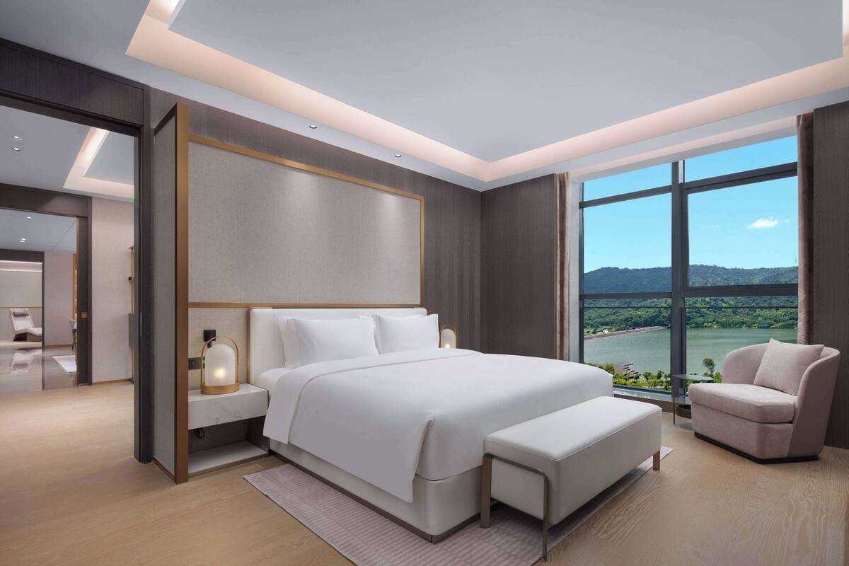 Ein modernes Schlafzimmer mit einem großen Bett, Nachttischen, einer Leselampe und Blick auf einen See durch große Fenster.