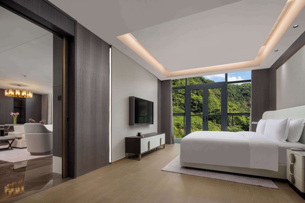 Ein modernes Schlafzimmer mit einem großen Bett, einem an der Wand montierten Fernseher und Blick auf Grün durch große Fenster.