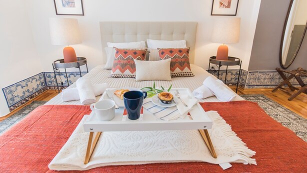 Una cama bien hecha con una bandeja de desayuno, que incluye un croissant y una taza, en una habitación con alfombra roja y almohadas estampadas.
