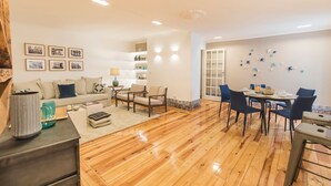 Un salón amplio con piso de madera, un conjunto de sofás, una mesa de comedor con sillas y una cocina al fondo.
