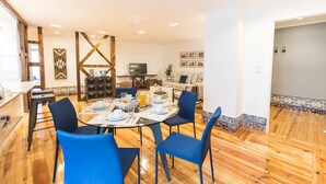 Un comedor con una mesa redonda dispuesta para cuatro personas, rodeada de sillas azules. Se observa un aparador de madera con cajones y un televisor al fondo.
