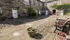 Un edificio de piedra con múltiples ventanas y puertas, un patio empedrado con un banco y plantas en macetas.