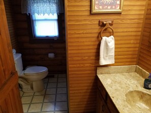 Ein Badezimmer mit Holzwänden, einer Toilette, einem Waschbecken und einem Fenster mit Spitzenvorhängen.