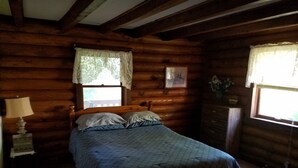 Une chambre à coucher dans un chalet en rondins, avec un lit, une table de chevet, une commode et une fenêtre ornée de rideaux en dentelle.