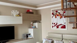 A modern living room with a sofa, a dining table, a TV, and a kitchen area with a staircase.