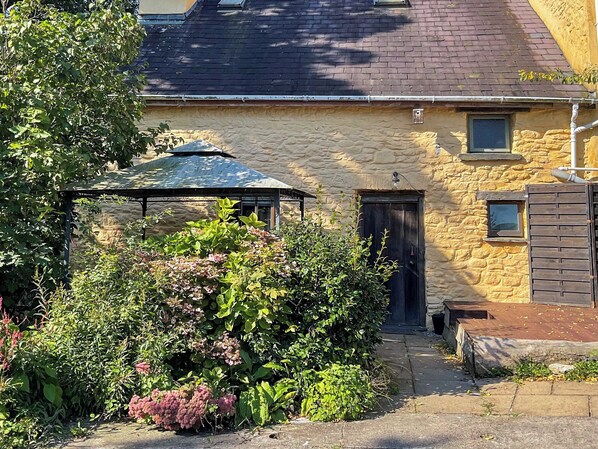 Un bâtiment en pierre avec un toit sombre, un espace extérieur couvert et un jardin où poussent différentes espèces de plantes.