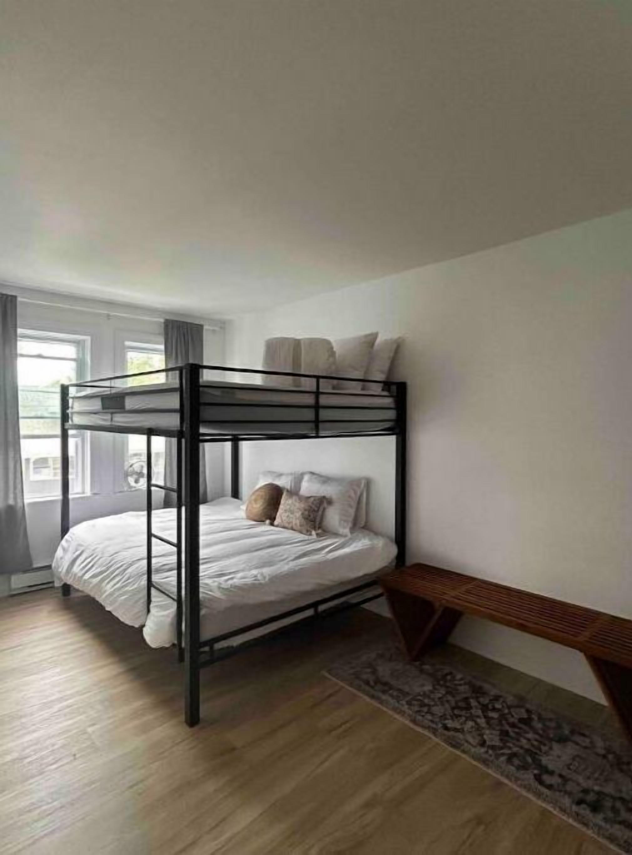 A bunk bed with a wooden frame and white bedding, a small bench, and a patterned rug in a room with wooden flooring and a window with curtains.
