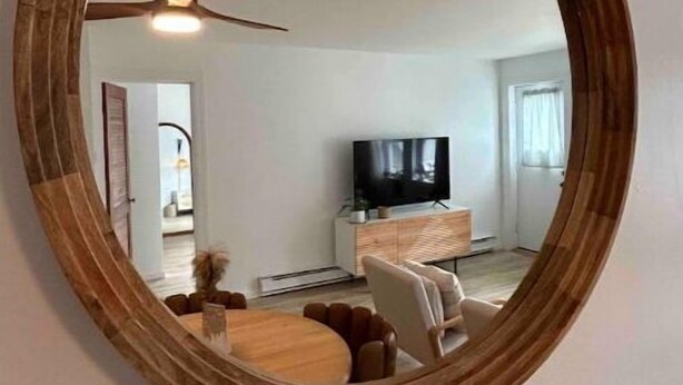 Un miroir rond, doté d’un cadre en bois, reflète un salon avec un ventilateur de plafond, un téléviseur et un coin repas.