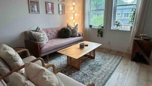 Un salon comprenant un canapé, un fauteuil et une table basse. Des tableaux encadrés ornent le mur, et une fenêtre est agrémentée de rideaux.